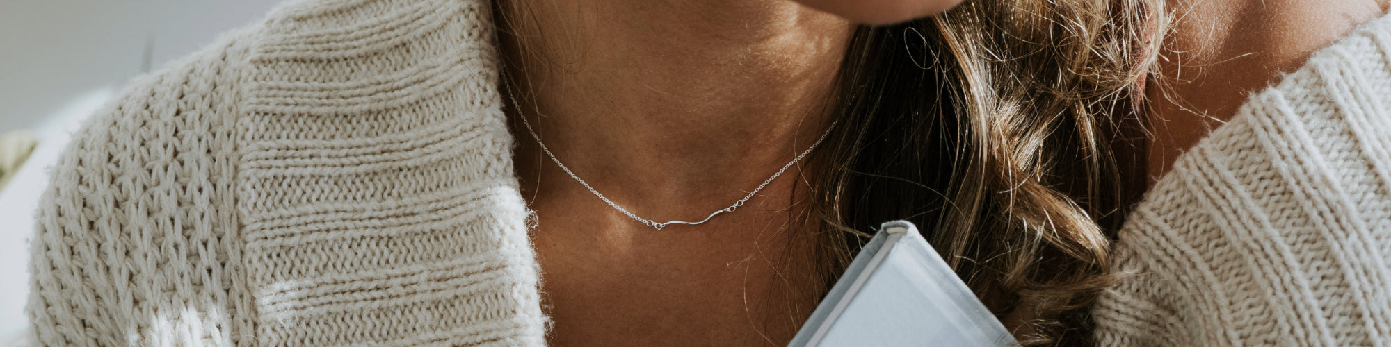 Close-up image of a woman wearing a delicate chain necklace with a small, round pendant, set against her sweater and blouse.