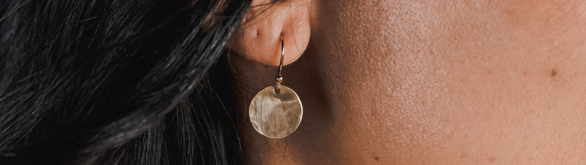 Close-up of a woman's ear wearing a round, metallic earring, with her black hair partially visible.