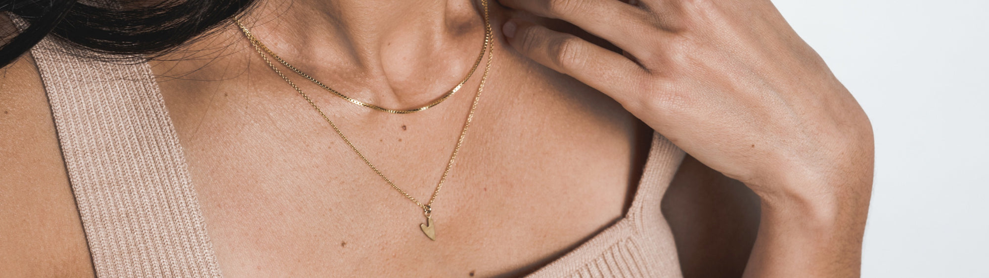 Close-up of a woman wearing a beige top and a delicate gold necklace with a pendant, touching her neck gently.