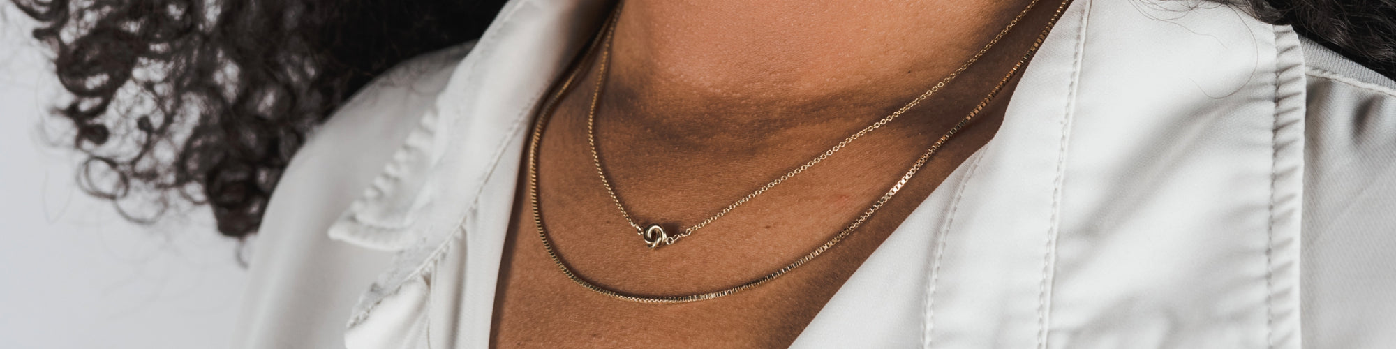 Close-up of a woman wearing a beige top and a delicate gold necklace with a pendant, touching her neck gently.