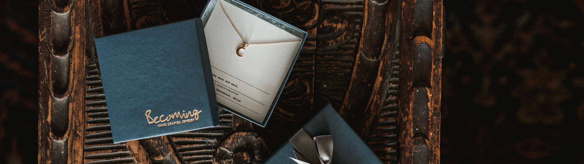 A delicate crescent moon necklace in a gift box labeled "Becoming," placed on a wooden surface alongside another closed gift box.