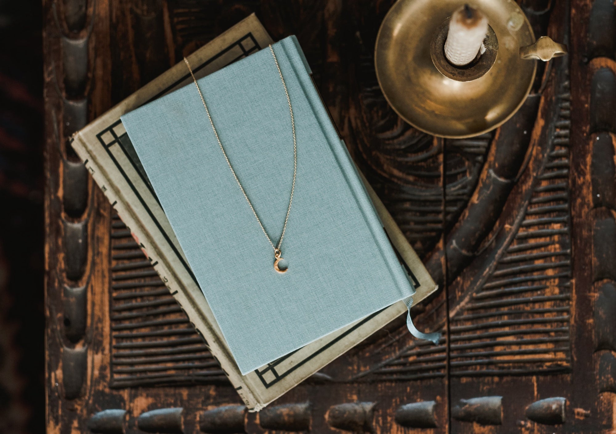 A blue hardcover book with a pendant necklace resting on it is placed on a decorative wooden surface, beside a brass candle holder.
