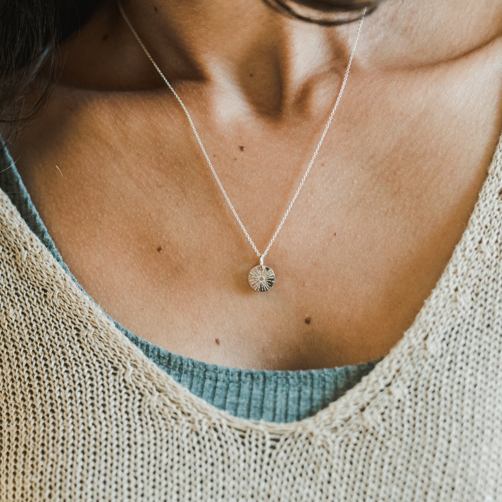 A person wearing the "Be The Light Necklace" by Becoming Jewelry, paired with a light blue top and a beige knitted sweater.