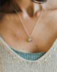 A person wearing the "Be The Light Necklace" by Becoming Jewelry, paired with a light blue top and a beige knitted sweater.