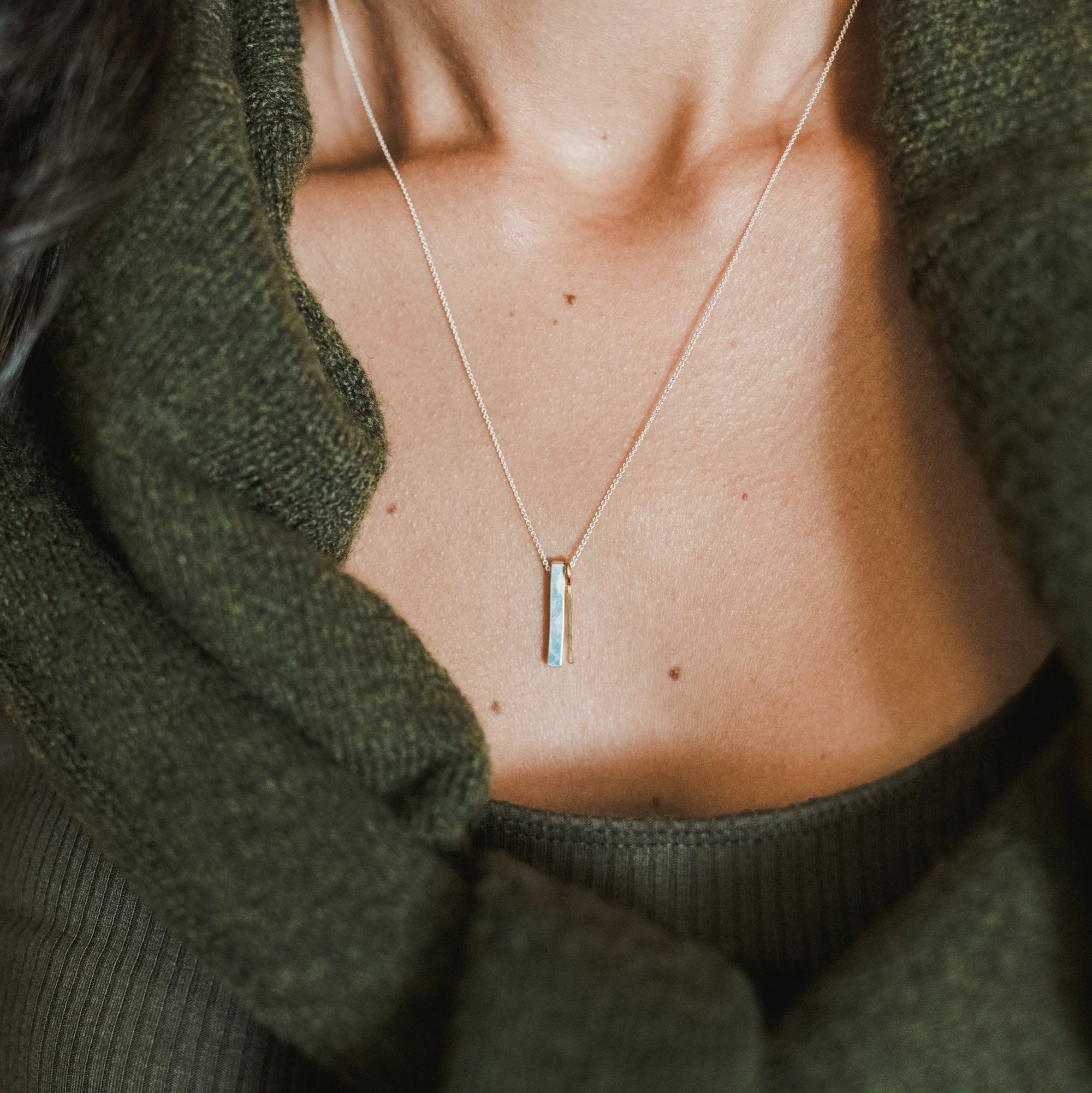 Close-up of a person wearing the Through Thick & Thin Necklace by Becoming Jewelry. This best-selling, simple, thin necklace with a small vertical pendant makes the perfect gift. The person is dressed in a green sweater and a dark green top underneath.