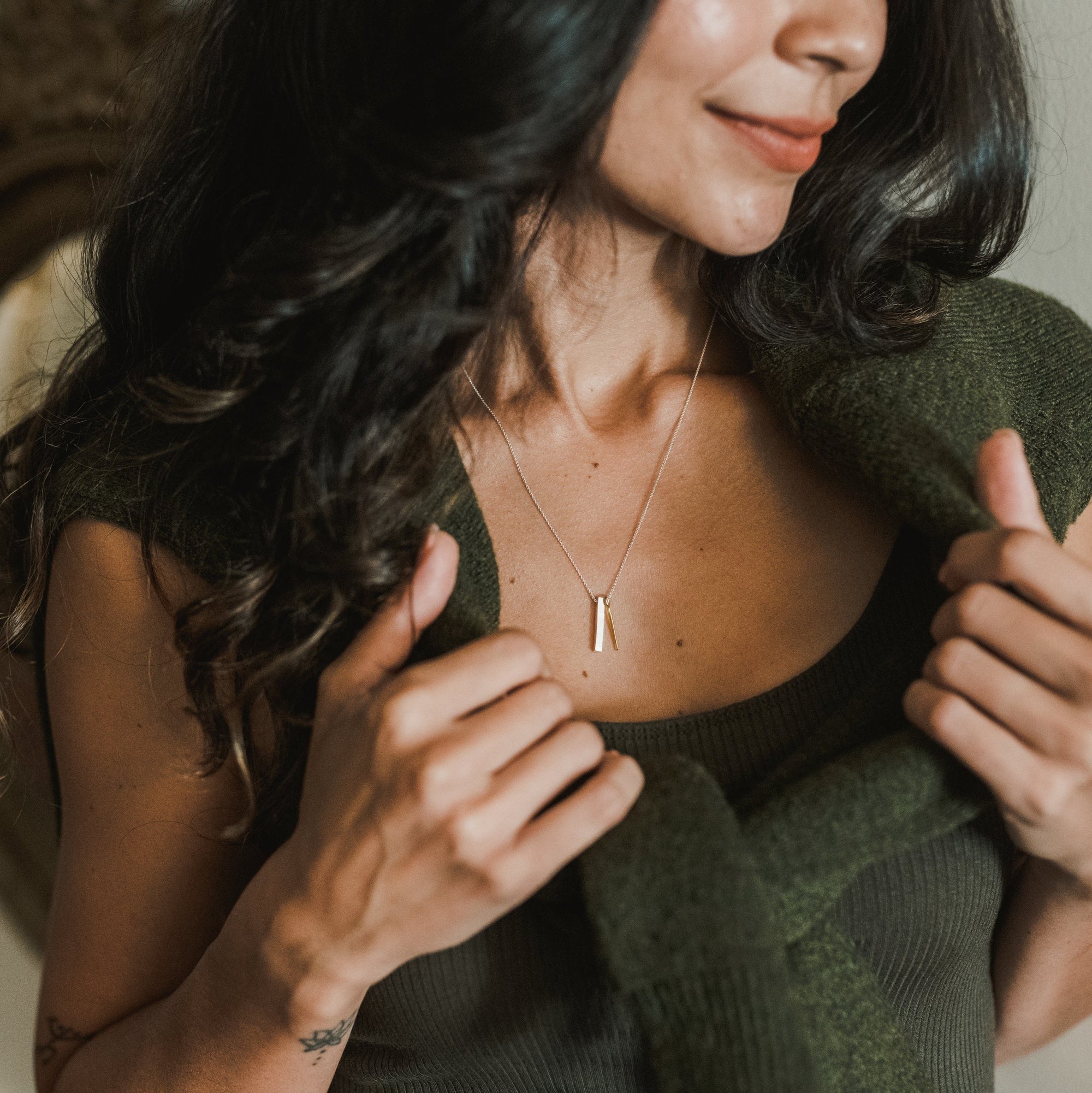 Close-up of a person with long, dark hair wearing a green top and Becoming Jewelry's best-selling Through Thick & Thin Necklace, holding a green sweater draped over their shoulders.