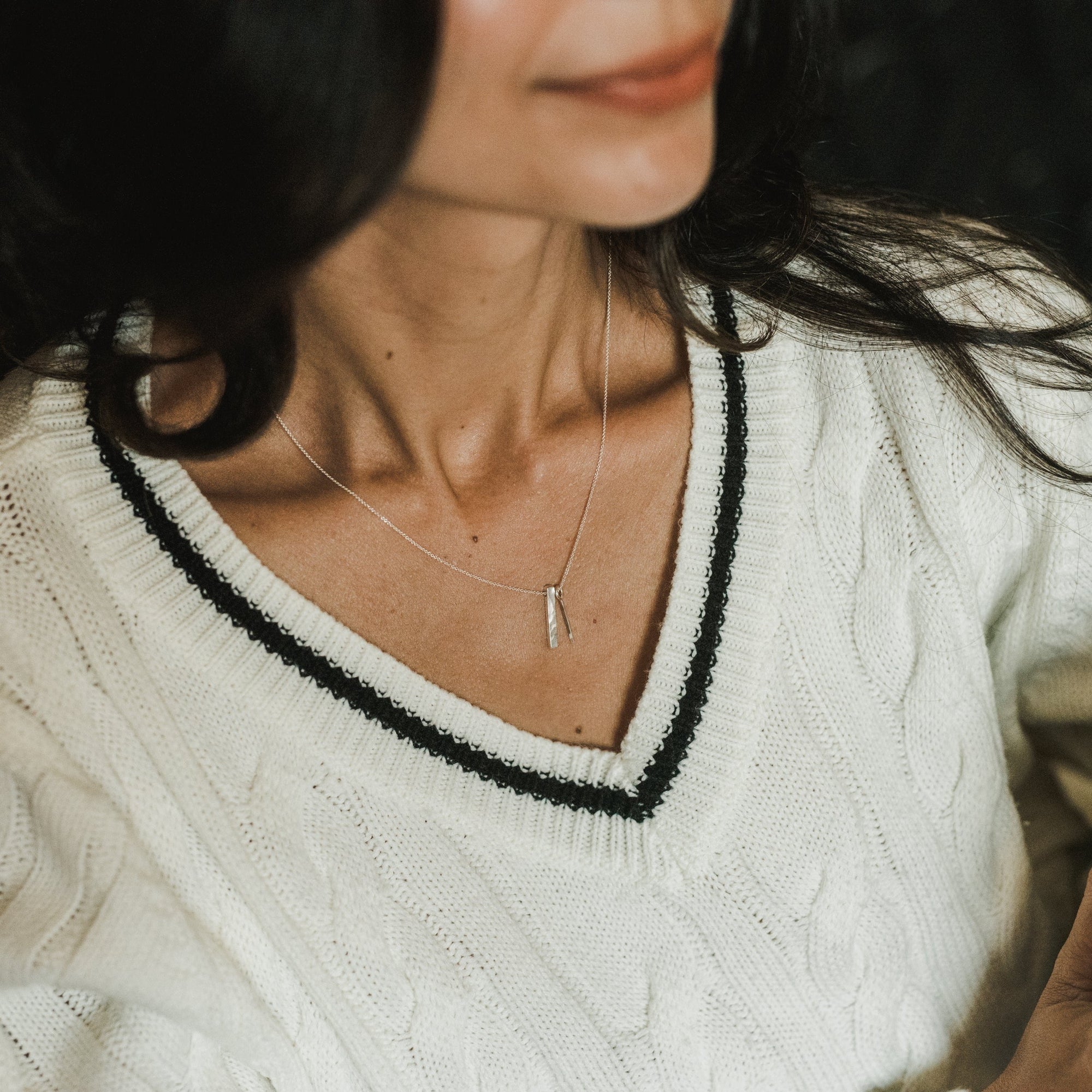Close-up of a person wearing a white cable-knit sweater with a black-trimmed V-neck and the delicate silver "Through Thick & Thin Necklace" by Becoming Jewelry. Their face is partially visible but out of focus, making it the perfect gift for someone special.
