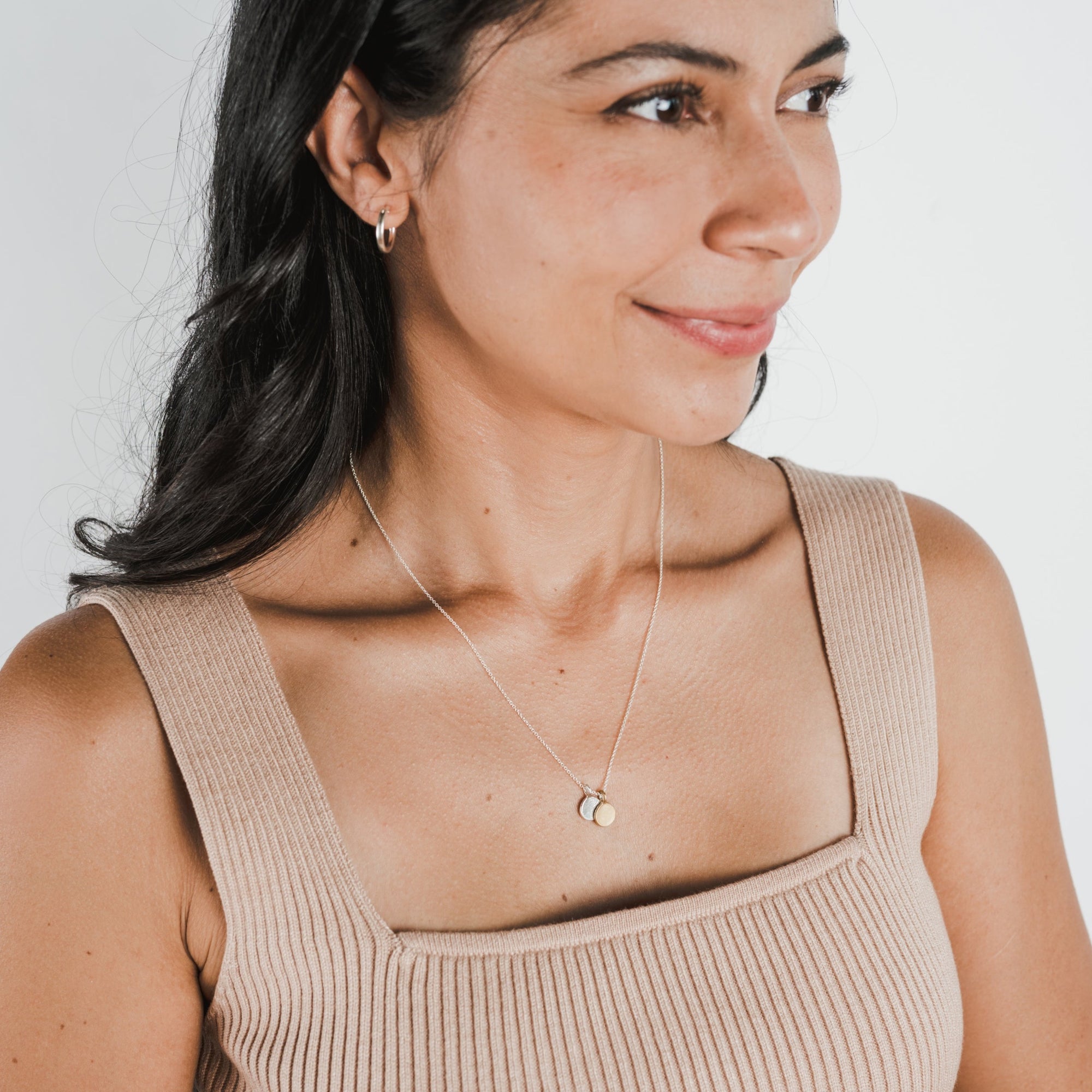A woman with long dark hair, wearing a beige sleeveless top, small hoop earrings, a Becoming Jewelry "Count My Blessings" necklace with a round pendant, smiles and looks to her right against a plain light background. Her charming gift of gold drop discs adds an extra touch of elegance to her style.