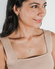 A woman with long dark hair, wearing a beige sleeveless top, small hoop earrings, a Becoming Jewelry "Count My Blessings" necklace with a round pendant, smiles and looks to her right against a plain light background. Her charming gift of gold drop discs adds an extra touch of elegance to her style.
