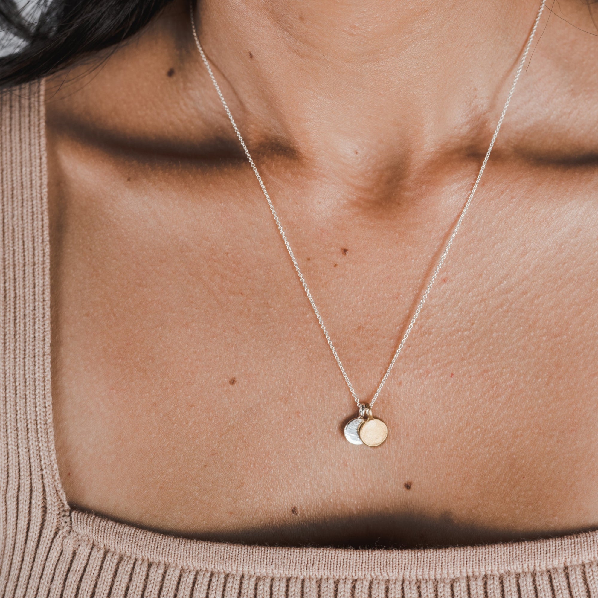 An individual is wearing the Count My Blessings Necklace by Becoming Jewelry, showcasing a thin, silver chain with two small, silver drop disc pendants. They are dressed in a beige, ribbed tank top.