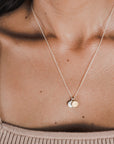 An individual is wearing the Count My Blessings Necklace by Becoming Jewelry, showcasing a thin, silver chain with two small, silver drop disc pendants. They are dressed in a beige, ribbed tank top.