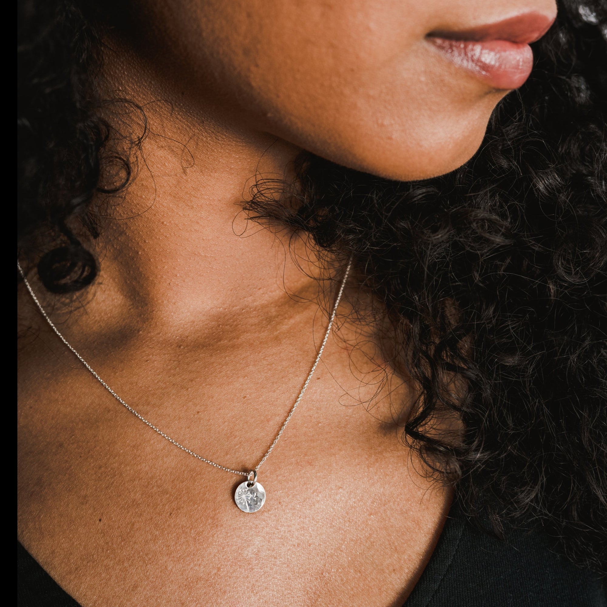 A close-up reveals a person with curly hair wearing the Dandelion Wishes Necklace by Becoming Jewelry—an elegant silver chain adorned with a round pendant featuring inspirational words.
