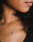 A close-up reveals a person with curly hair wearing the Dandelion Wishes Necklace by Becoming Jewelry—an elegant silver chain adorned with a round pendant featuring inspirational words.