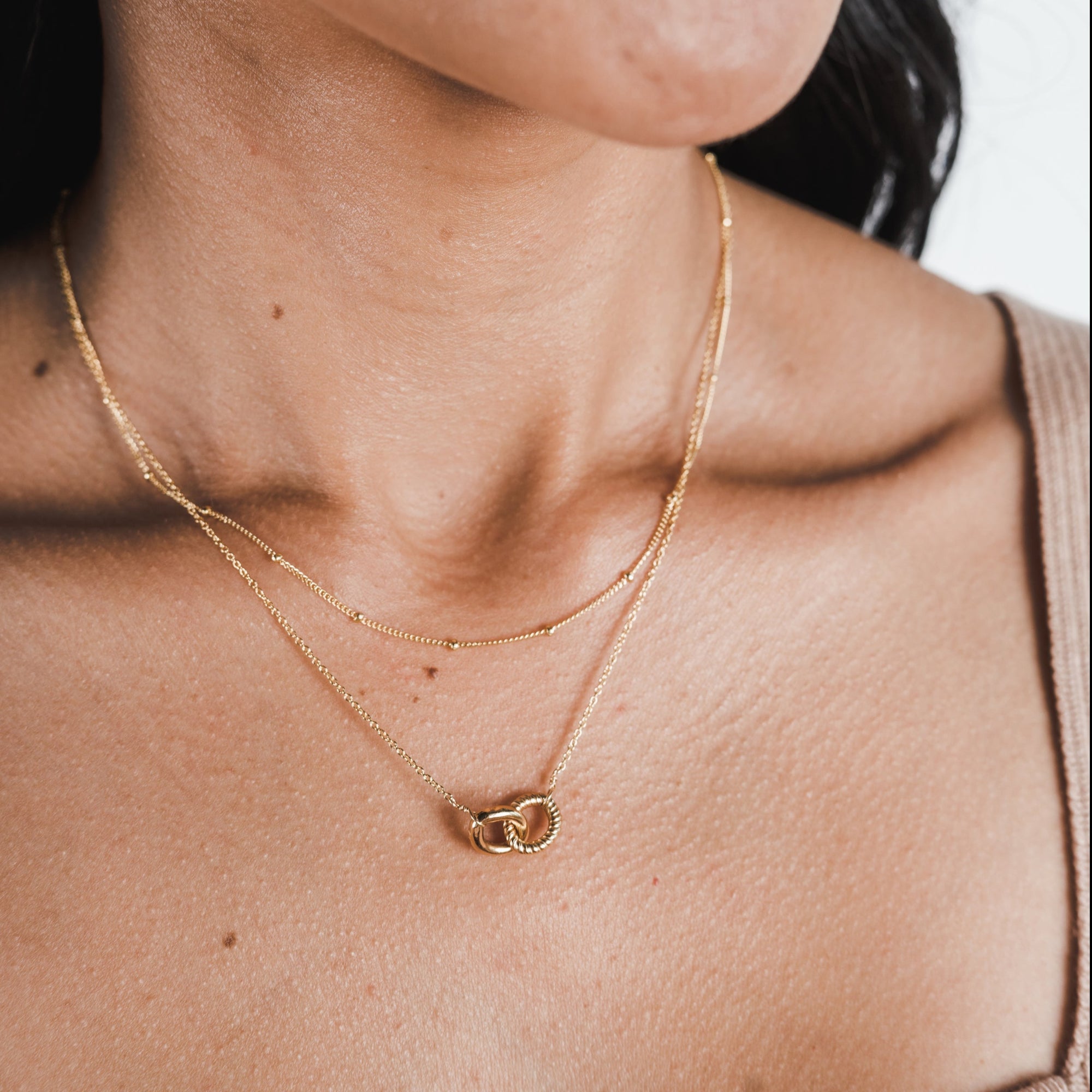 Close-up of a person wearing two gold necklaces. One is an everyday necklace with a small delicate chain, and the other is the Satellite Chain Necklace from Becoming Jewelry, featuring a circular intertwined pendant. Only the lower part of the face is visible, showcasing how effortlessly these pieces can layer with other necklaces.