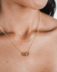 Close-up of a person wearing two gold necklaces. One is an everyday necklace with a small delicate chain, and the other is the Satellite Chain Necklace from Becoming Jewelry, featuring a circular intertwined pendant. Only the lower part of the face is visible, showcasing how effortlessly these pieces can layer with other necklaces.