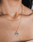 Close-up of a person wearing two gold necklaces from Becoming Jewelry, with The Sea Necklace's pendant featuring an embossed design of a wave pattern, reminiscent of the ocean. A light-colored top is also partially visible.