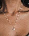 Close-up of a person's neck adorned with two delicate silver necklaces, featuring Becoming Jewelry's Figaro Chain Necklace paired with another necklace showcasing a small padlock pendant. The layered look is enhanced by the Figaro chain, adding to the delicate texture. The neutral background highlights the jewelry beautifully.