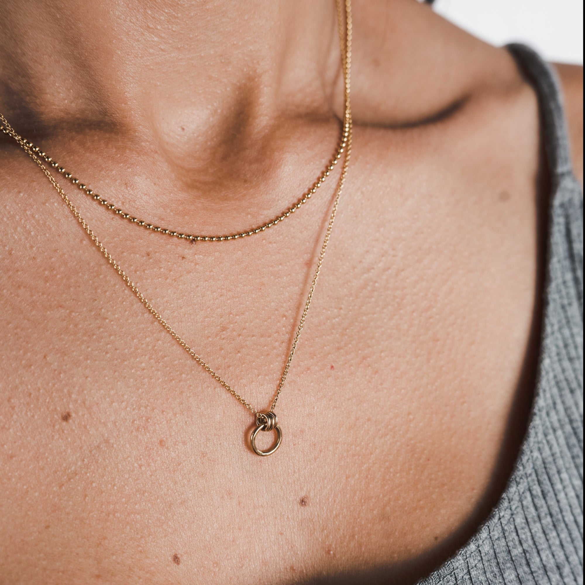 Close-up of a person's neck wearing two gold necklaces from Becoming Jewelry. One features a small chain, while the other showcases the Family Circle Necklace with its circular pendant, symbolizing strength and love. The person is dressed in a ribbed grey top, making these pieces the perfect go-to gift.