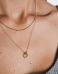 Close-up of a person's neck wearing two gold necklaces from Becoming Jewelry. One features a small chain, while the other showcases the Family Circle Necklace with its circular pendant, symbolizing strength and love. The person is dressed in a ribbed grey top, making these pieces the perfect go-to gift.