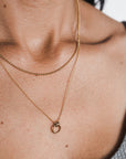 A close-up shot captures a person's neck beautifully adorned with two delicate, layered gold necklaces from Becoming Jewelry. One is the Bead Chain Necklace, featuring a thin chain that adds a subtle elegance to the look. The other necklace showcases a small circular pendant, adding texture and charm to the ensemble. The person complements the necklaces perfectly with a gray sleeveless top.