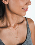 Close-up of a woman wearing a gray top, showcasing gold hoop earrings and two layered necklaces—one is the Bead Chain Necklace from Becoming Jewelry and the other features a small ring pendant, adding texture to her elegant ensemble.