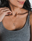 A person wearing a gray ribbed tank top showcases the Surrounded By Love Necklace from Becoming Jewelry, featuring two interlocking rings as a meaningful reminder of connection, and has a floral tattoo on their wrist.