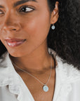 A person with curly hair wears a white blouse, a Becoming Jewelry Strong Women Necklace, and matching earrings, facing slightly to the side.