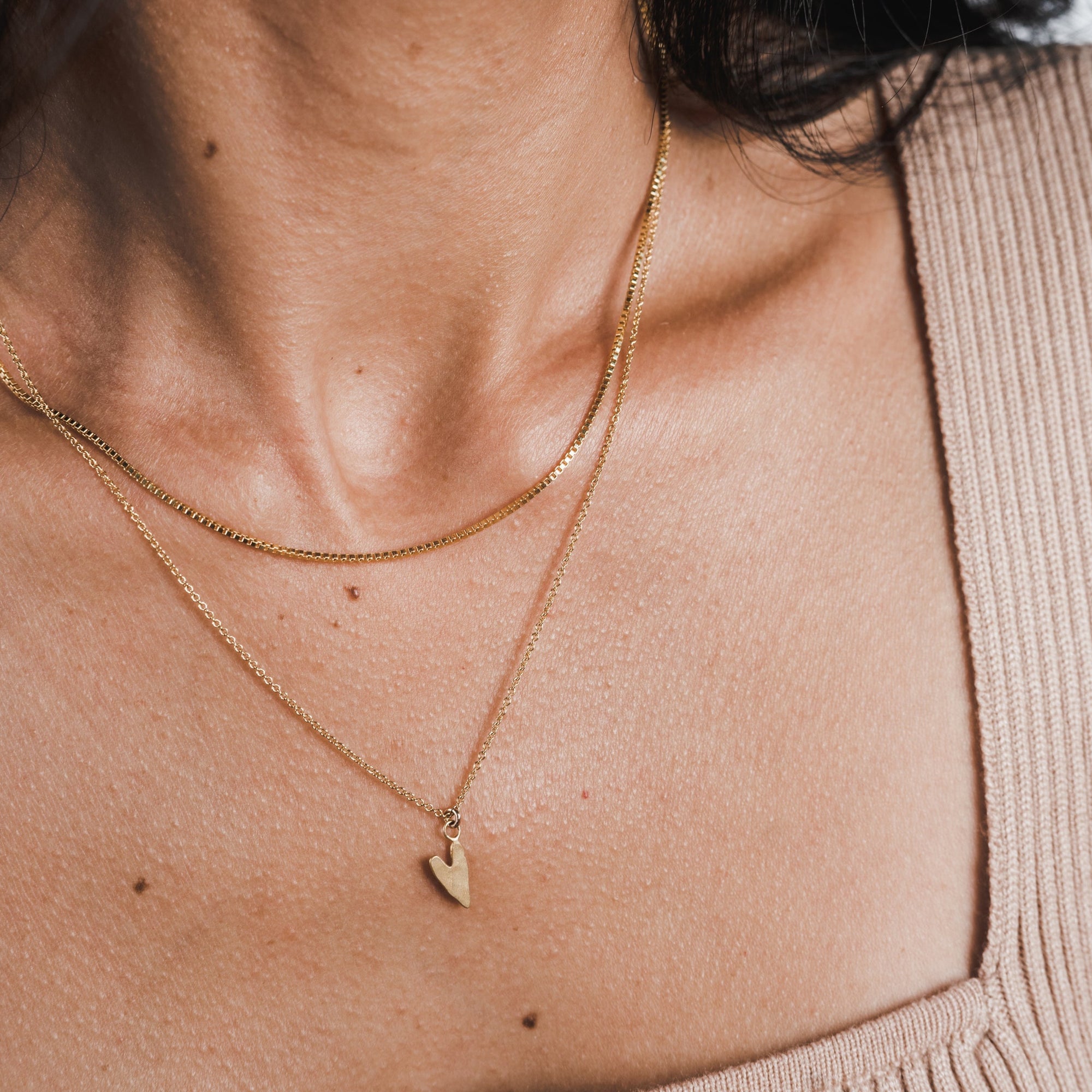 Close-up of a woman's neck adorned with two gold necklaces: one featuring a precious heart charm and the other, the Love Deeply Necklace by Becoming Jewelry. She is dressed in a beige sleeveless top.