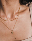 Close-up of a woman's neck adorned with two gold necklaces: one featuring a precious heart charm and the other, the Love Deeply Necklace by Becoming Jewelry. She is dressed in a beige sleeveless top.
