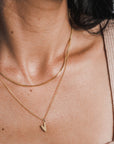 Close-up of a person's neck wearing two lightweight gold necklaces from Becoming Jewelry, including the Box Chain Necklace with a classic look featuring a small heart-shaped pendant, paired with a beige sleeveless top.
