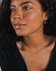 A person with curly hair wearing a black top, a thin gold Bead Chain Necklace from Becoming Jewelry, and a single hoop earring looks to the side against a plain background.