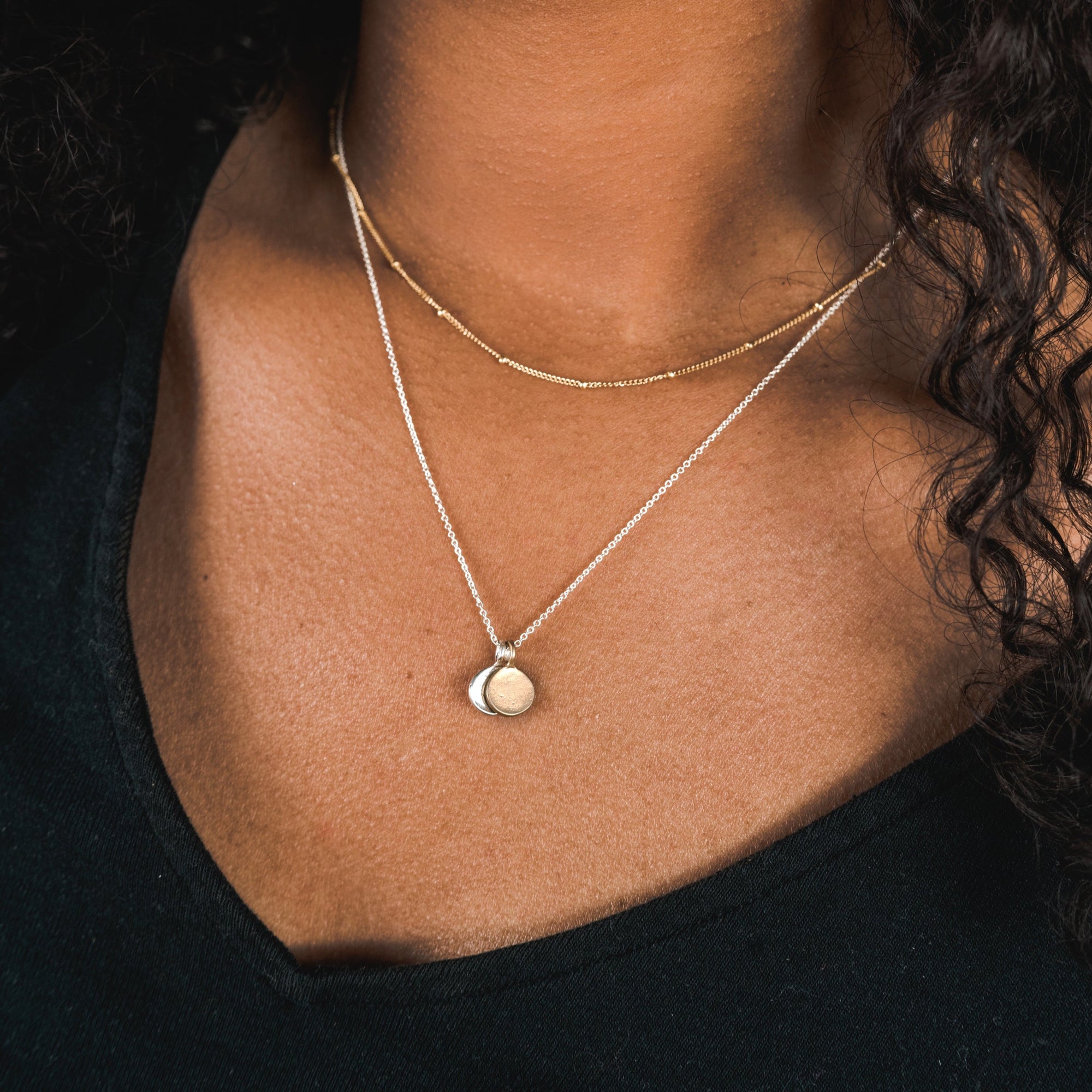 A close-up of a person wearing a black top, adorned with two gold necklaces from Becoming Jewelry; one is the elegant Satellite Chain Necklace, and the other features a coin-shaped pendant, perfect for layering to achieve an elegant everyday look.
