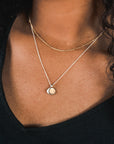 A close-up of a person wearing a black top, adorned with two gold necklaces from Becoming Jewelry; one is the elegant Satellite Chain Necklace, and the other features a coin-shaped pendant, perfect for layering to achieve an elegant everyday look.