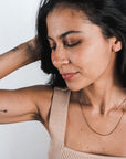 A woman with long dark hair in a beige sleeveless top touches her hair. She wears minimalist jewelry from Becoming Jewelry, including a Box Chain Necklace, and has a tattoo on her upper arm, completing her classic look.