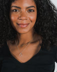 A person with long curly hair and a nose ring smiles slightly. They are wearing a black top and layered necklaces, one delicate with a circular pendant alongside the classic Curb Chain Necklace by Becoming Jewelry.