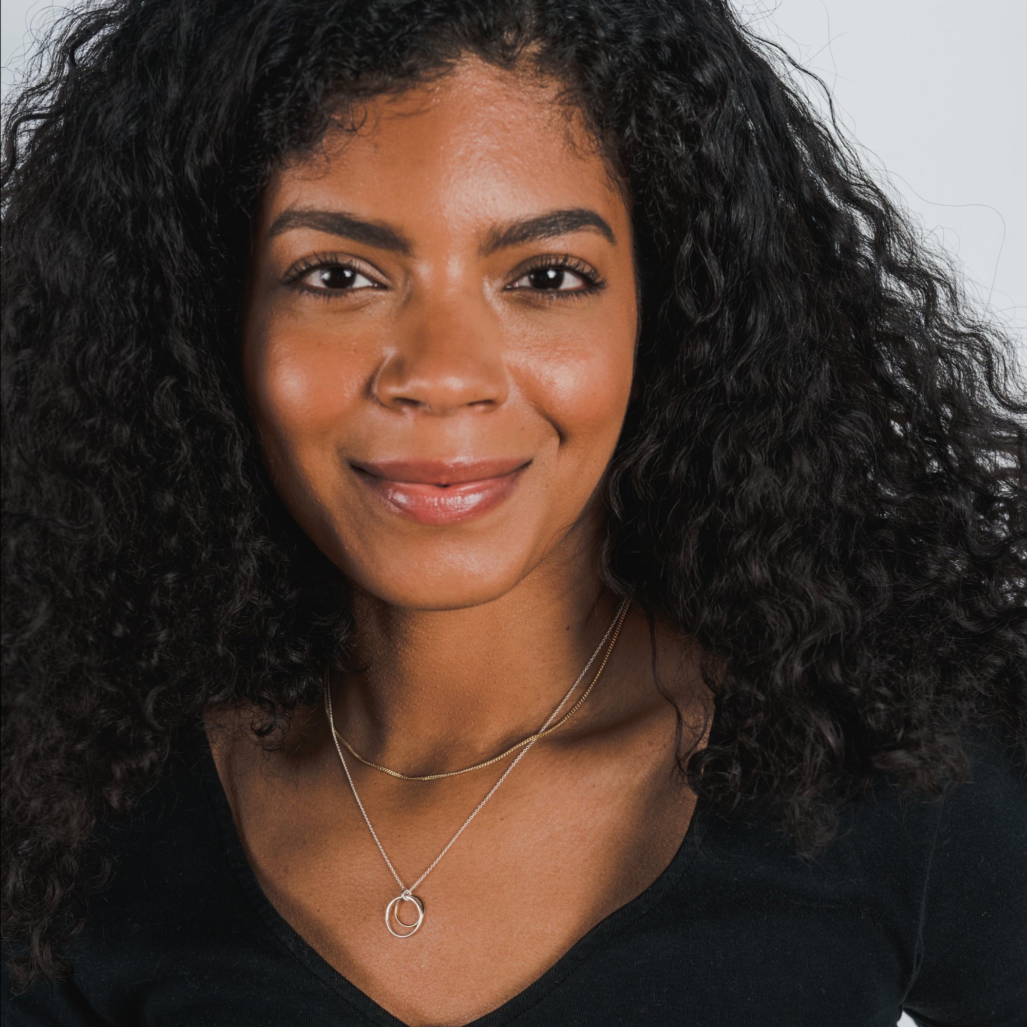 A woman with curly black hair wearing a black top and the "Surrounded By Love Necklace" by Becoming Jewelry smiles towards the camera, perhaps showcasing it as a meaningful reminder, or considering it as a gift to someone special.