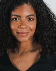A woman with curly black hair wearing a black top and the "Surrounded By Love Necklace" by Becoming Jewelry smiles towards the camera, perhaps showcasing it as a meaningful reminder, or considering it as a gift to someone special.