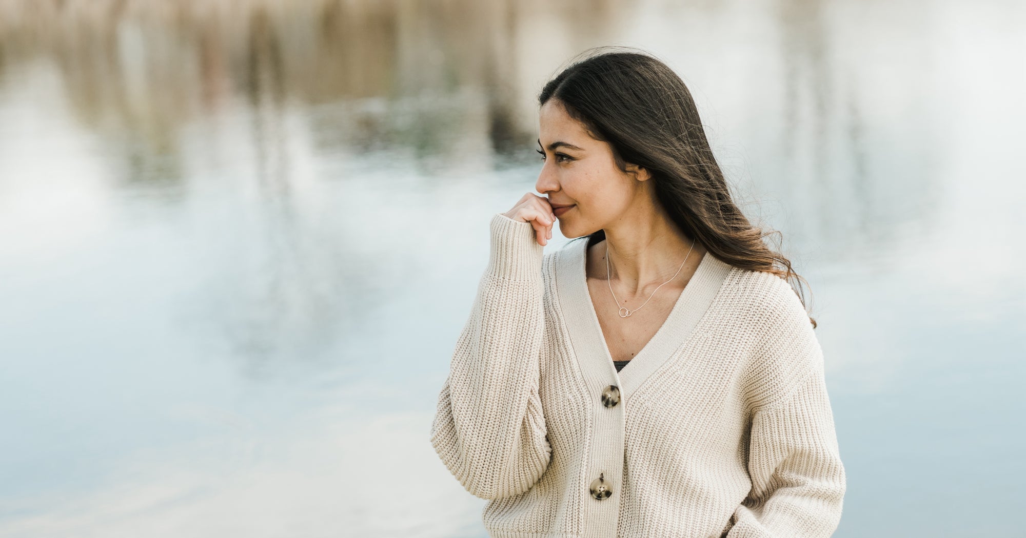 Woman in a beige sweater standing thoughtfully beside a body of water.