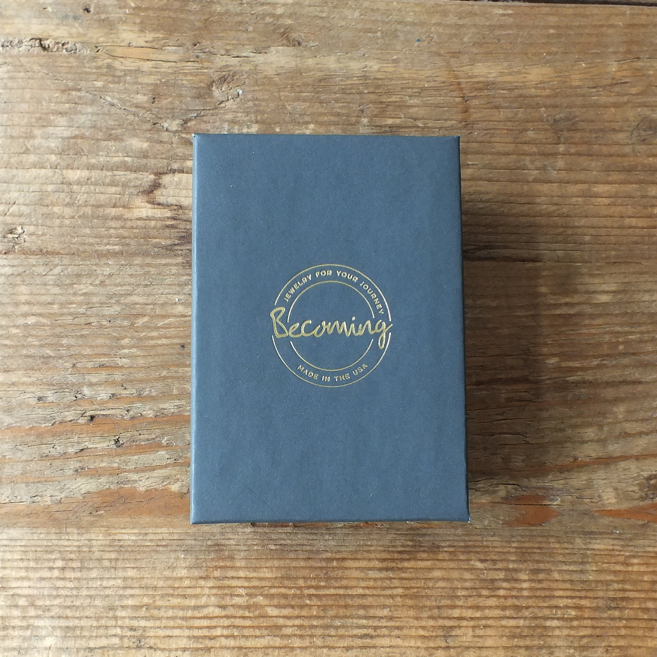 A closed black box labeled Becoming Jewelry with gold text rests elegantly on a wooden surface, echoing the refined craftsmanship of the Hammered Disc Drop Earrings, tiny.