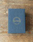 A closed black box labeled Becoming Jewelry with gold text rests elegantly on a wooden surface, echoing the refined craftsmanship of the Hammered Disc Drop Earrings, tiny.