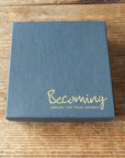 On a wooden surface lies a square, dark gray box adorned with the gold-etched Becoming Jewelry for Your Journey, carrying an aura of love and mindfulness, housing the May You Be Happy Necklace by Becoming Jewelry.