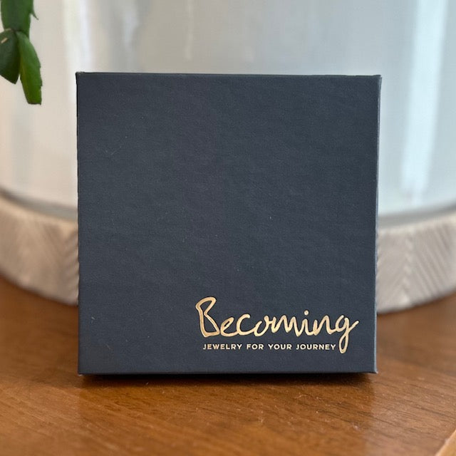 On a wooden surface sits a navy blue box with gold text reading Becoming Jewelry and Jewelry for your journey. Inside, the Little Things Necklace is held, offering a beaded chain as the perfect meaningful gift and daily reminder of your personal path.