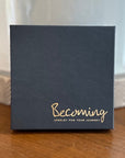 On a wooden surface sits a navy blue box with gold text reading Becoming Jewelry and Jewelry for your journey. Inside, the Little Things Necklace is held, offering a beaded chain as the perfect meaningful gift and daily reminder of your personal path.