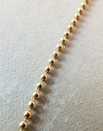 Close-up image of a gold Bead Chain Necklace by Becoming Jewelry, each bead connected in a linear sequence, layered gracefully on a textured beige surface.