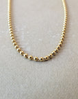 A close-up image of the Becoming Jewelry Bead Chain Necklace, elegantly arranged in a U-shape on a light beige surface.