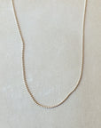 A thin, delicate Bead Chain Necklace by Becoming Jewelry is showcased against a neutral background, exuding an elegant layered texture.
