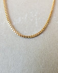 Close-up image of a lightweight gold Box Chain Necklace by Becoming Jewelry, featuring small square links, displayed on a light beige surface, offering a classic look.