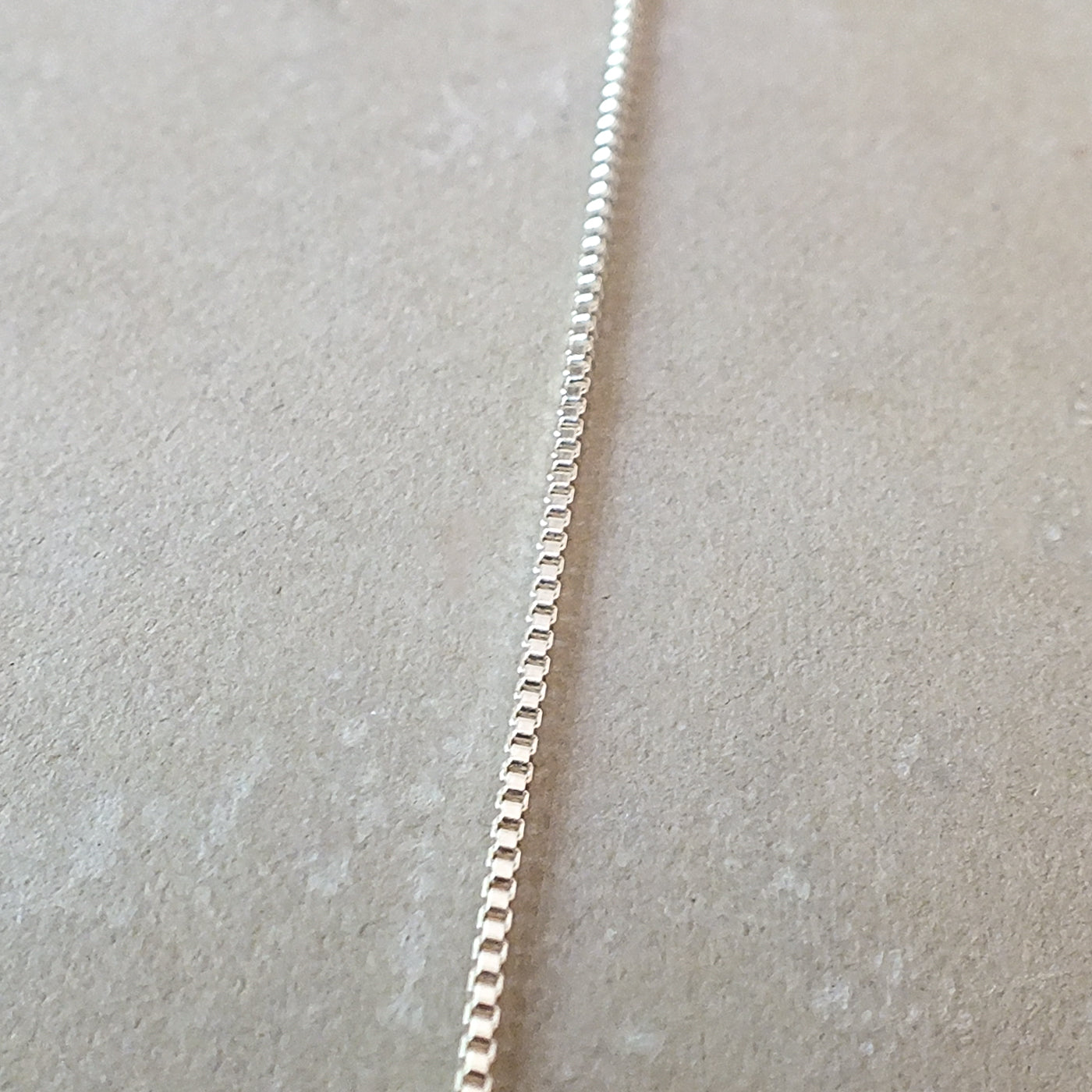 A close-up of a delicate Becoming Jewelry sterling silver box chain necklace lying on a textured surface.