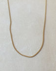A simple, delicate **Curb Chain Necklace** from **Becoming Jewelry** is laid out against a plain, light-colored background, perfect for layering with other classic curb chain or layered necklaces.