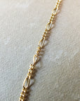 Close-up image of the Becoming Jewelry Figaro Chain Necklace with small, interlocking links placed on a beige surface, enhancing its delicate texture.